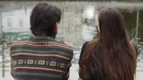 Two adults seated at urban pond sharing quiet conversation with gentle hand gestures under overcast daylight