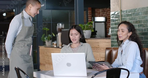 Diverse female coworkers at cafe table with laptop, tablet, gesturing to server in apron speaking