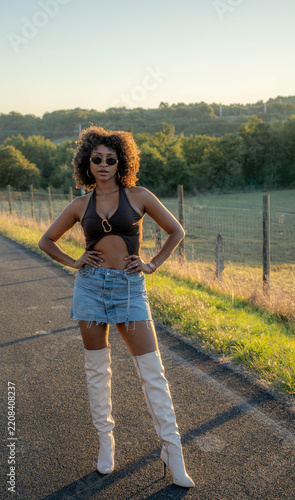 Black woman standing on quiet rural road during warm sunset light, wearing sunglasses, black halter top, denim skirt, and white boots, with hands on hips against peaceful countryside fields.