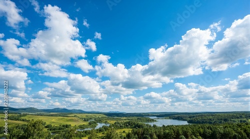 Vast sky holds fluffy white clouds above green rolling hills. River winds through forested valley below. Sunlight glows warmly over distant tree line