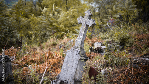 Old leaning stone cross gravestone in an overgrown cemetery. Weathered abandoned tombstone surrounded by dry grass and weeds in autumn. Spooky historical graveyard background