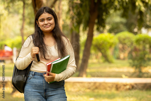 Portrait of indian young girl college student with backpack and notebooks standing and smiling in park