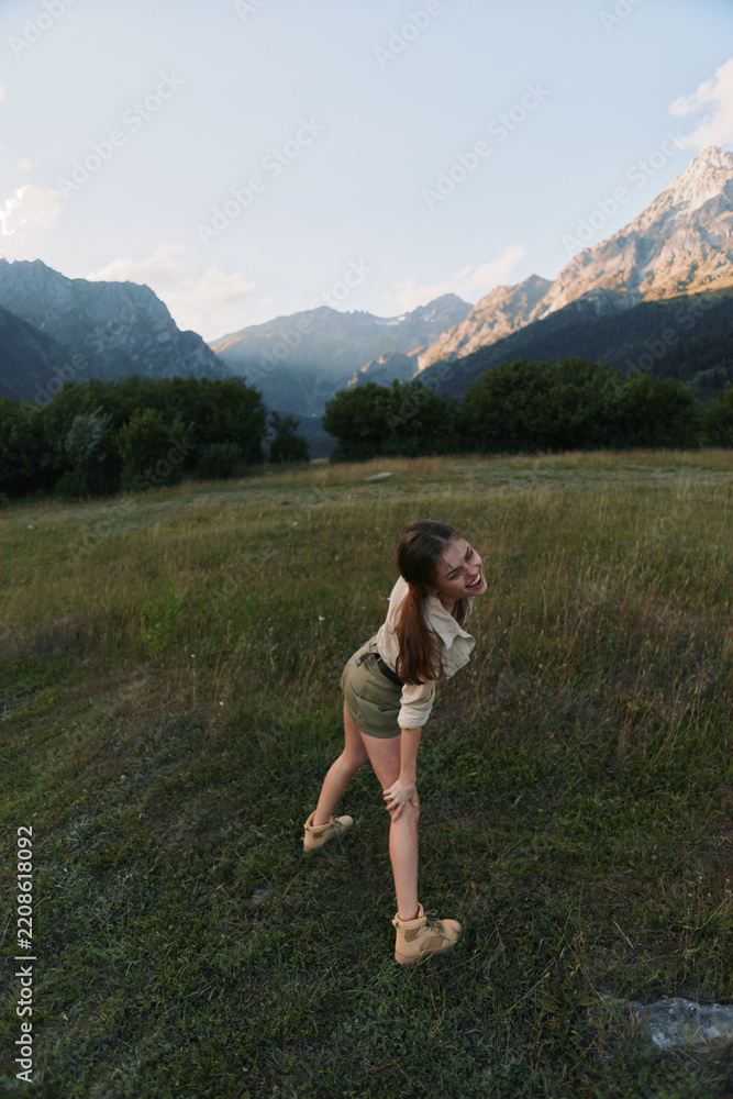 Obraz premium Young woman in a light hiking outfit stands in a wide grassy field with towering mountains in the background, enjoying a carefree outdoor moment.