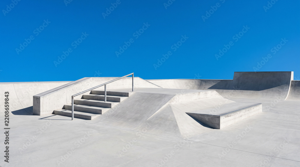 Obraz premium A pristine, modern concrete skatepark featuring stairs, handrails, and ramps under a vivid clear blue sky. Ideal for urban lifestyle and extreme sports content.