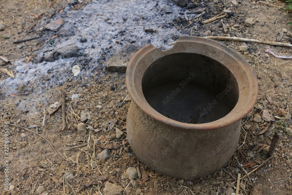 Fototapeta premium Earth-toned pot sits beside smoldering ashes. Dry ground holds scattered pebbles and dried grass. Pot shows signs of use and age, its rim worn. Scene evokes quiet aftermath of outdoor cooking