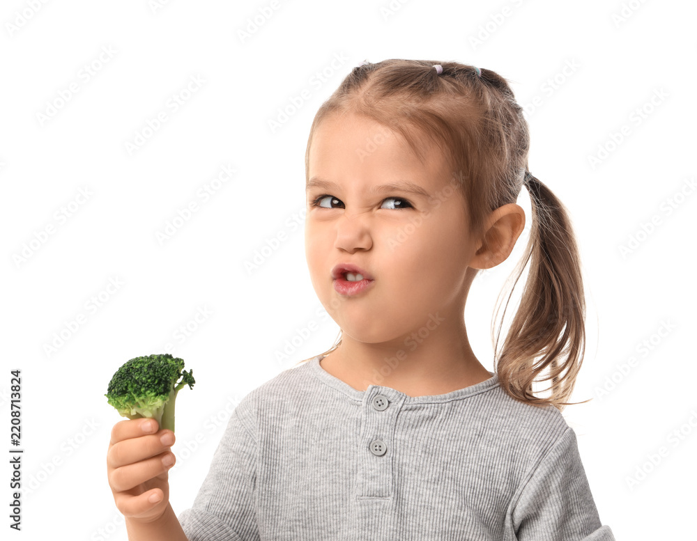 Fototapeta premium Healthy eating habits and picky eater concept, little girl making a disgusted face while holding a piece of fresh green broccoli isolated