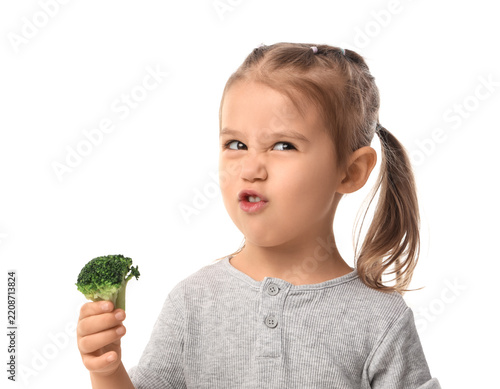 Healthy eating habits and picky eater concept, little girl making a disgusted face while holding a piece of fresh green broccoli isolated