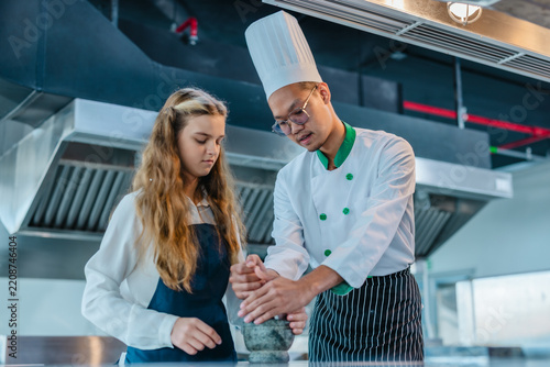 Asian chef teaching teen culinary intern to use mortar and pestle in a professional kitchen