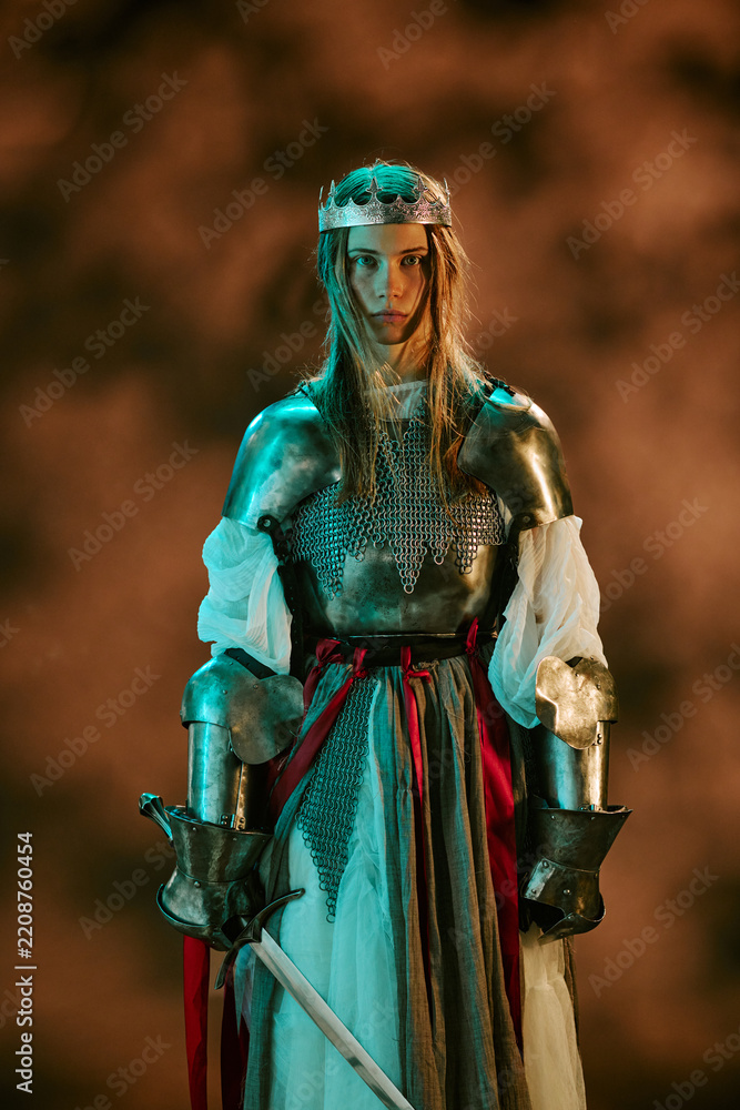 Obraz premium Portrait of young adult Caucasian woman wearing armor and crown holding sword representing leadership and strength. Media suitable for historical film promotion, gaming campaigns, vertical