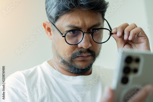 Middle-aged Asian man lifting his glasses to read text on his smartphone