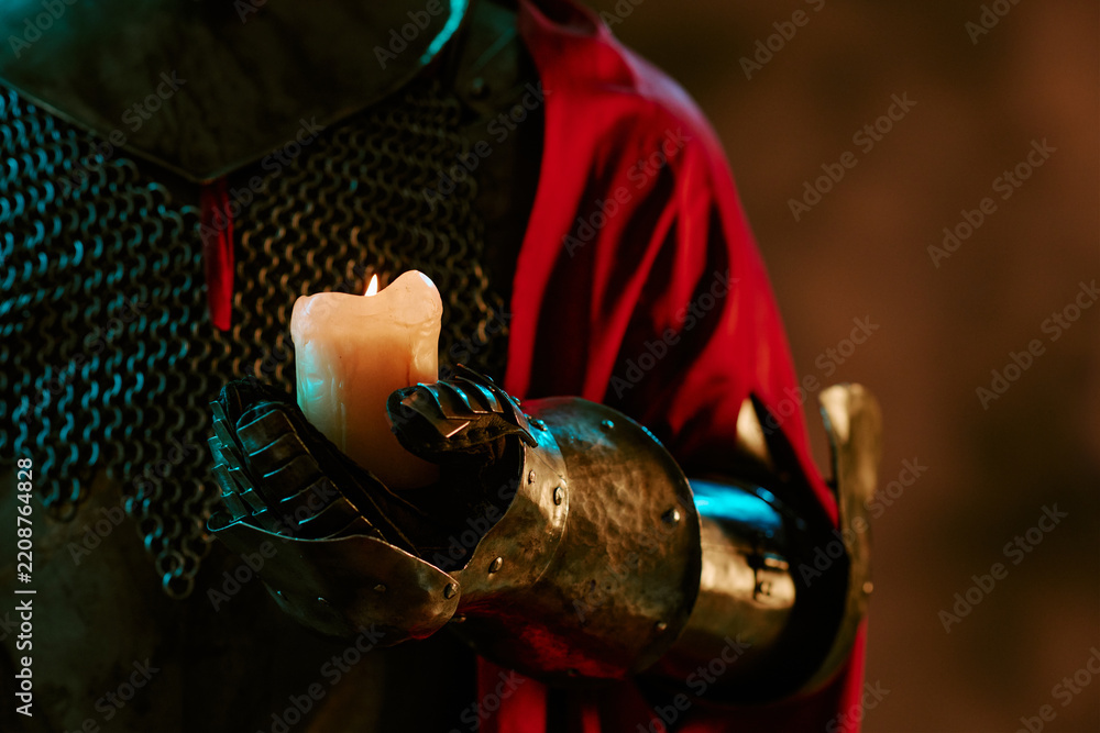 Obraz premium Medieval knight wearing metal armor gauntlet holding burning candle, representing hope and guidance. Useful for fantasy game promotion, book covers, and theatrical posters