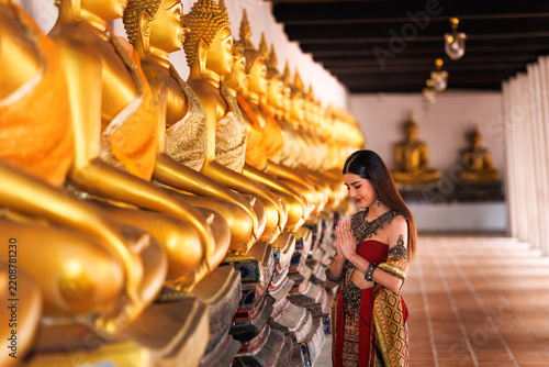 Graceful Asian Woman In Traditional Thai Silk Costume Performing Respectful Wai Prayer Beside Golden Buddha Statues In An Ancient Temple Cloister In Thailand