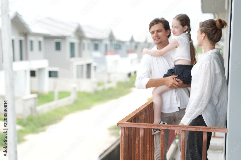 Fototapeta premium Young parents holding their child enjoy the view of the growing suburb from their balcony on the balcony of their new house overlooking the residential estate, shallow depth of field