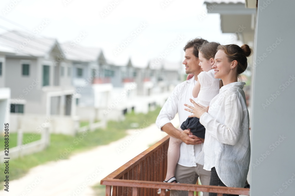 Fototapeta premium Happy young family with little daughter enjoy the view of the growing suburb from their balcony on the upper balcony of their new home above the neighborhood, authentic everyday scene