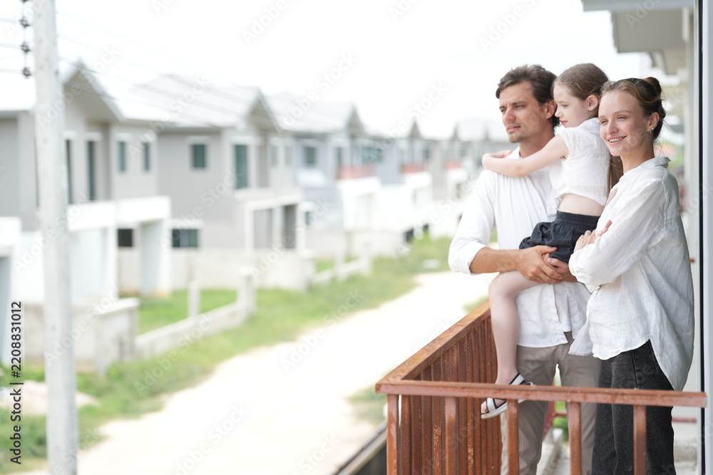 Fototapeta premium Caucasian family of three embracing enjoy the view of the growing suburb from their balcony standing on the terrace of a newly built suburban home, candid real moment