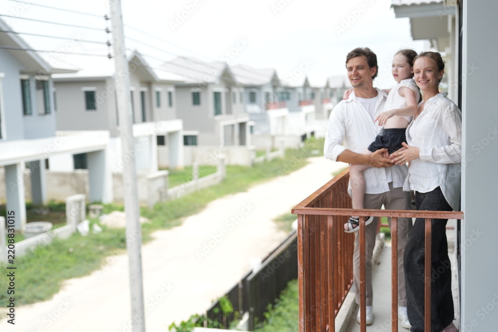 Fototapeta premium Happy young family with little daughter look out over the rows of new white houses from the railing on the upper balcony of their new home above the neighborhood in warm natural light