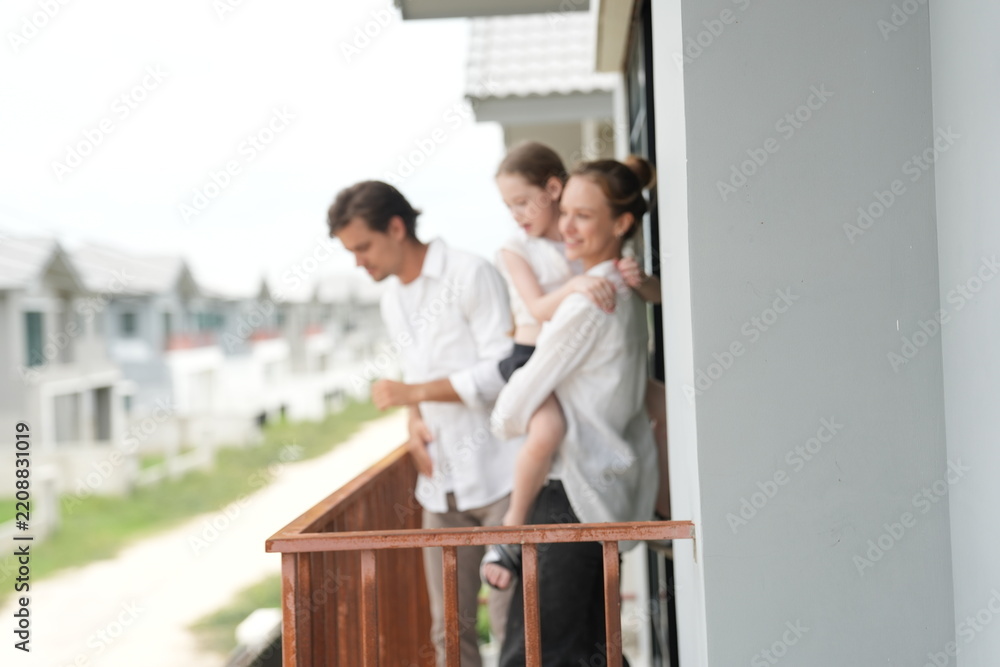 Fototapeta premium Happy young family with little daughter stand at the wooden railing admiring the neighborhood view standing on the terrace of a newly built suburban home on a bright sunny day