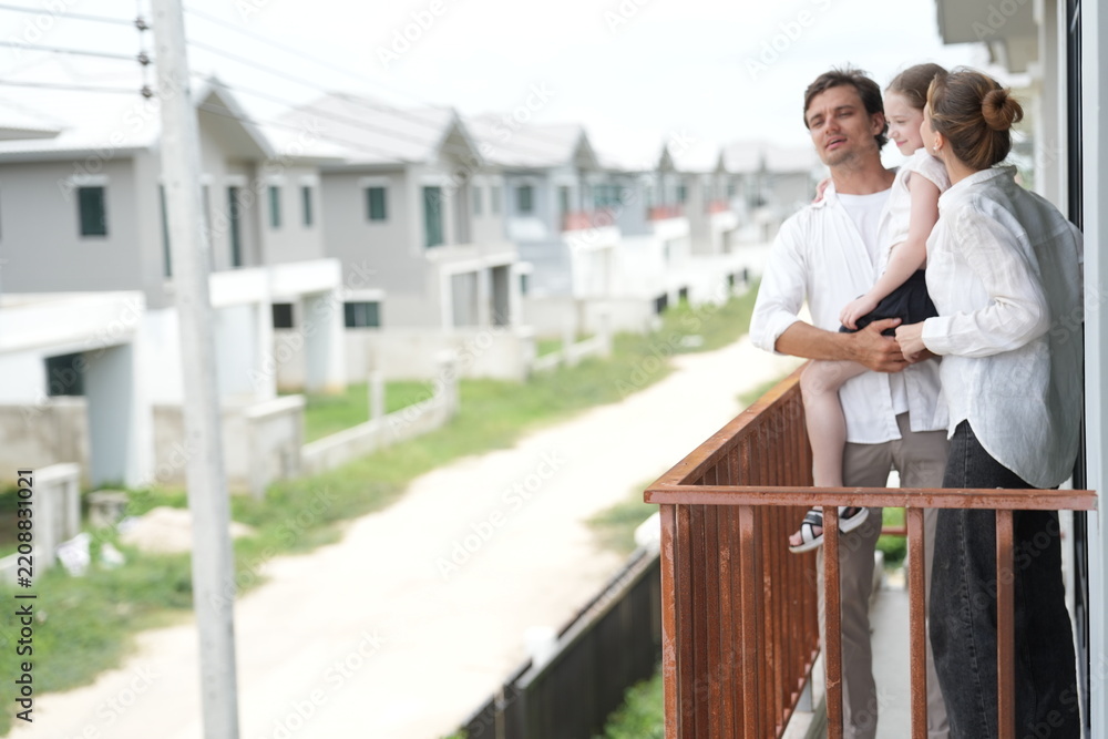 Fototapeta premium Caucasian family of three embracing look out over the rows of new white houses from the railing on the upper balcony of their new home above the neighborhood, shallow depth of field
