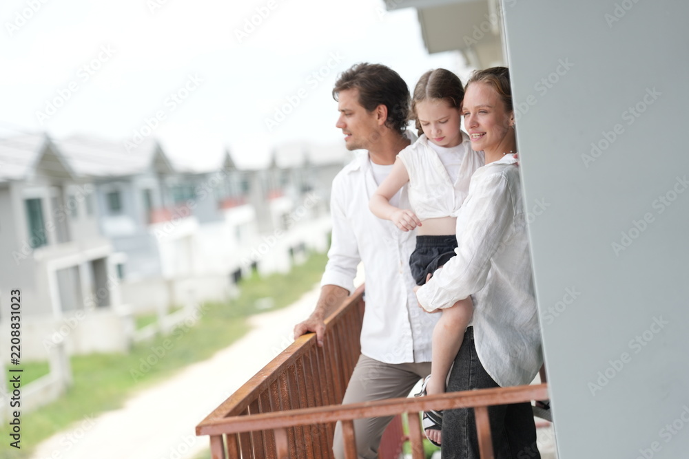 Fototapeta premium Caucasian family of three embracing enjoy the view of the growing suburb from their balcony on the balcony of their new house overlooking the residential estate, authentic everyday scene