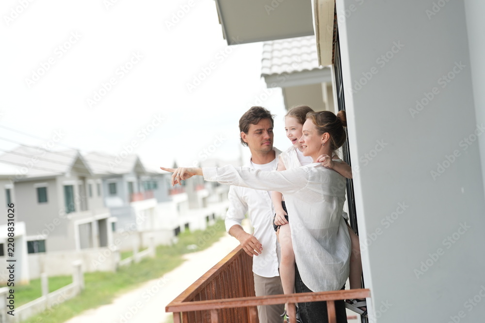 Fototapeta premium Young parents holding their child stand at the wooden railing admiring the neighborhood view standing on the terrace of a newly built suburban home, shallow depth of field