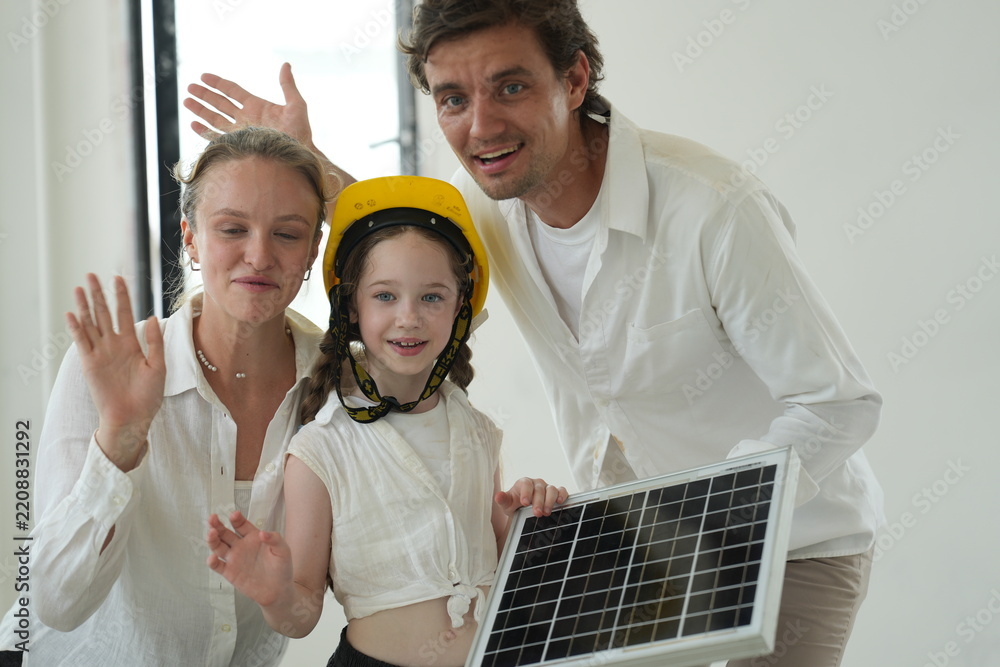 Obraz premium Young parents teaching their curious child kiss their daughter who proudly holds the photovoltaic module in the bright interior of their newly built home, shallow depth of field