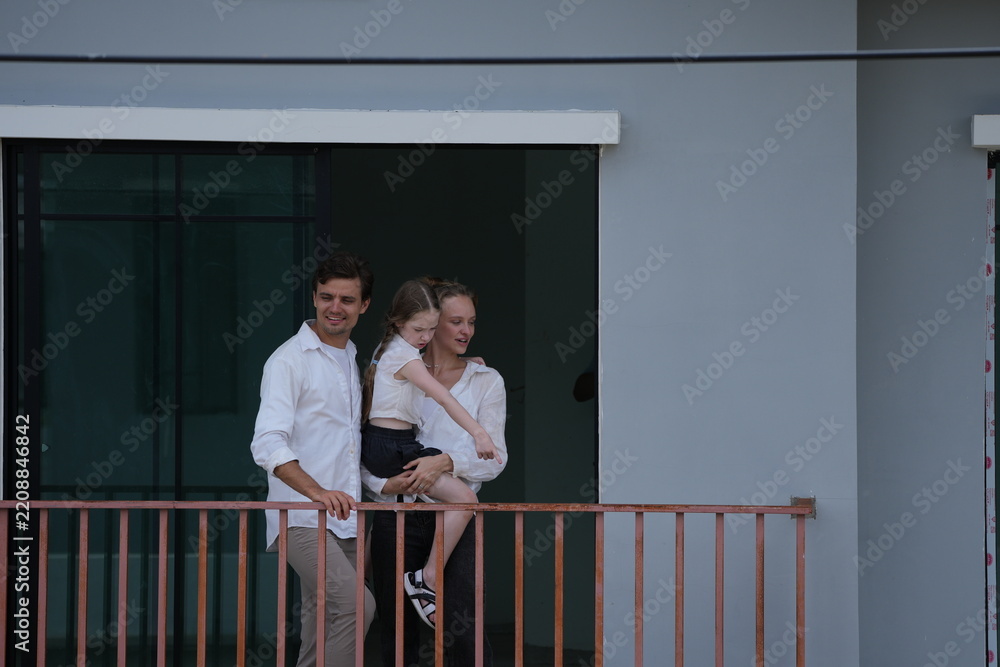 Obraz premium Young parents holding their child greet the neighborhood from the balcony with their daughter standing on the terrace of a newly built suburban home on a bright sunny day