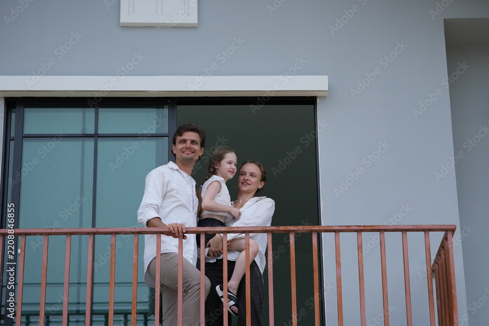 Obraz premium Happy young family with little daughter greet the neighborhood from the balcony with their daughter on the balcony of their new house overlooking the residential estate, candid real moment