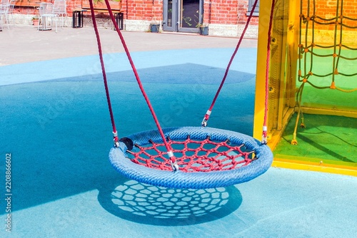 Colorful circular swing with red netting and blue ropes hangs in a sunlit playground, casting a shadow on the blue rubber floor.