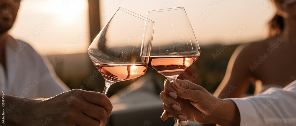 Obraz premium Romantic celebration moment as two people toast with elegant rosfilled glasses at sunset. Warm golden light glows through crystal, perfect for wine, lifestyle, and luxury dining campaigns.