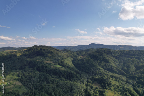 Panoramic drone view of vast green mountain ridges and pine forests under bright blue sky with white clouds