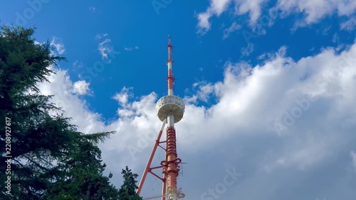 Telecommunications tower rising above trees under a bright blue sky with clouds. Infrastructure scene illustrating wireless communication, broadcasting, connectivity and modern technology.