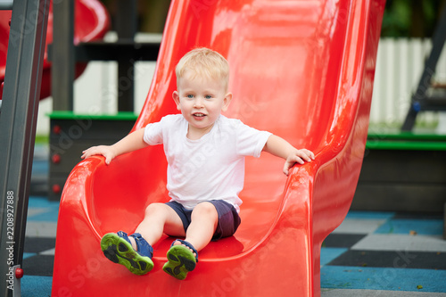 Obraz na plátně Child sliding down playground slide, holding sides and smiling, summer outfit, b