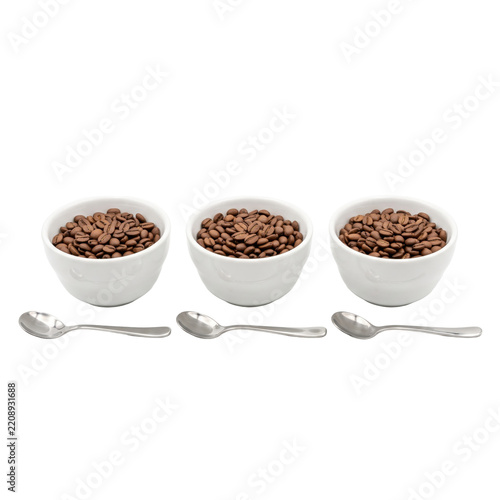 Three white ceramic bowls filled with roasted coffee beans and silver spoons isolated on white