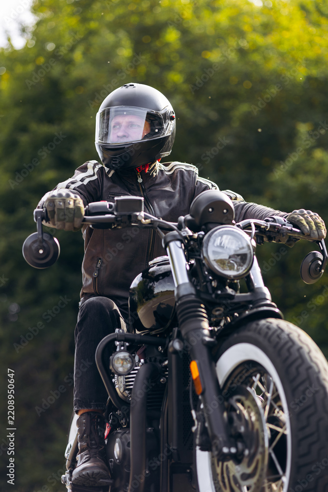 Obraz premium Biker wearing full-face helmet and leather jacket riding a vintage style motorcycle, looking forward on an outdoor adventure