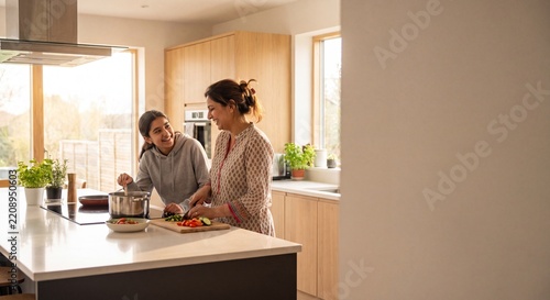 Happy Mother and Teenage Daughter Cooking Together in a Bright Modern Kitchen, Preparing Healthy Meal with Fresh Vegetables on a White Island