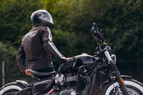 Motorcyclist wearing a black helmet and leather jacket, standing next to a classic black motorcycle with a blurred green background