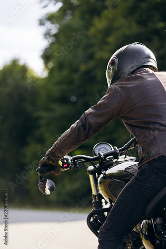 Person wearing leather jacket and helmet riding a classic motorcycle, traveling on an open road through nature