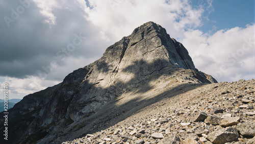Steep jagged outcrop of gray fractured rock rising against a partly cloudy sky, loose broken rocks and scree scattered across the barren talus slope in the foreground