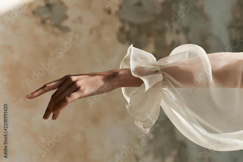 Graceful woman hand and arm extended in white organza sleeve against weathered beige textured background.