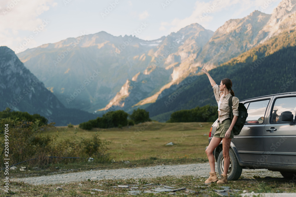 Obraz premium Young woman with a backpack stands beside a parked car in a mountain valley, raising her arm toward the sky as golden light hits rugged peaks, her expression curious and hopeful.