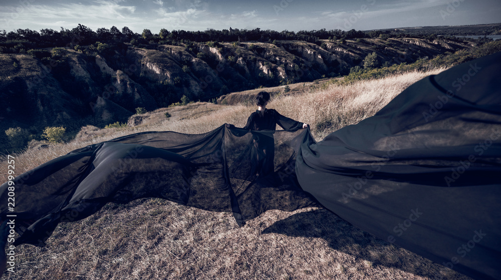Obraz premium Fearless figure extends black fabric like wings beneath dramatic sky with commanding energy. Personal power, freedom, cinematic fantasy concept