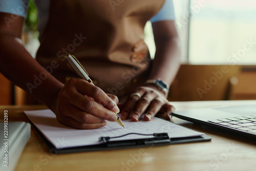 African American woman entrepreneur writing notes on clipboard near laptop representing small business management. Image suitable for accounting, retail planning and marketing