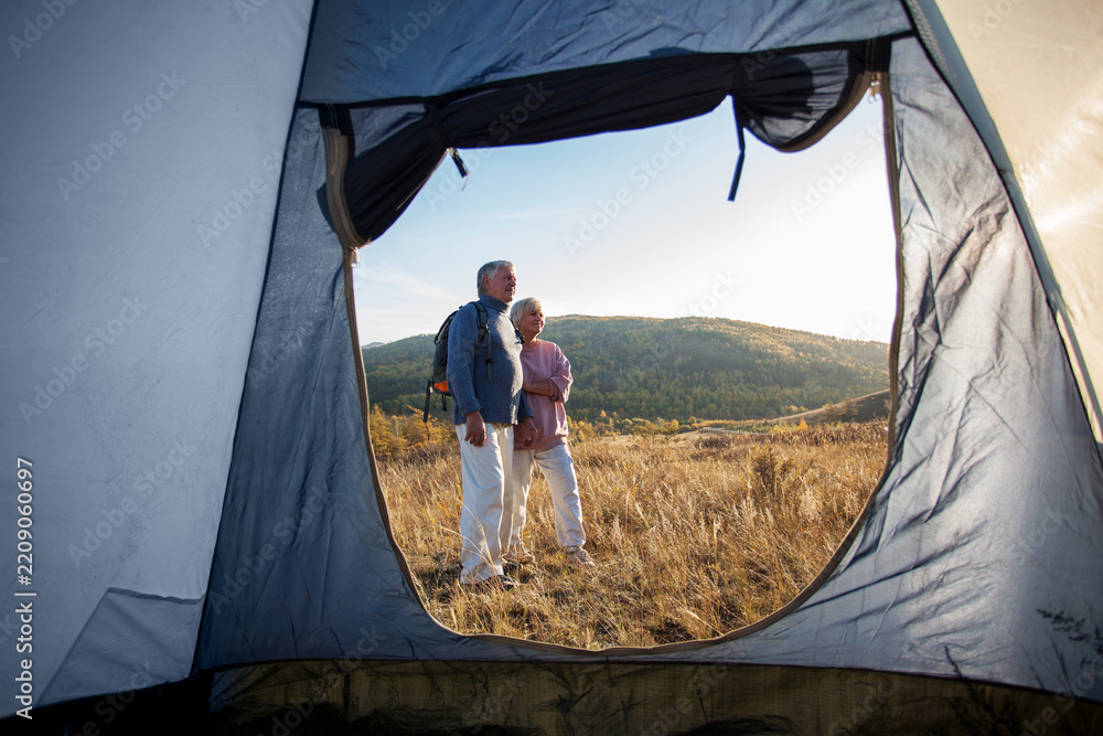 Obraz premium Older couple admires mountain views from grassy campsite, standing outside tent. Gray haired hikers wear backpacks and outdoor clothes, sharing peaceful moment in warm evening light.