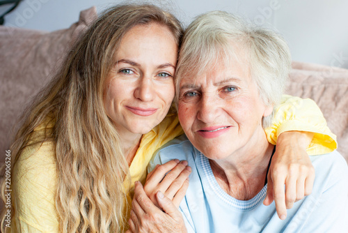 Young woman hugs elderly woman, her mom, on sofa, both smiling warmly at camera. Daughter embraces her older relative with arm around her shoulders, sharing close and affectionate family moment.