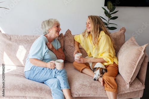 Older woman and younger woman laugh together on sofa, sharing relaxed visit over mugs of drink. Their small dog rests across younger womans lap in bright, welcoming living room, as women sit close