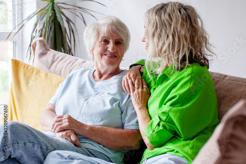 Older woman enjoys tender visit with younger woman seated close beside her. Smiling senior rests comfortably on sofa as her companion offers reassuring touch in welcoming living room.
