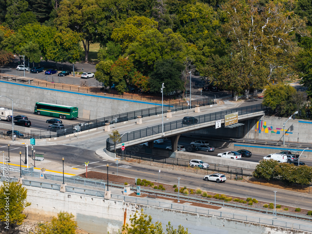 Obraz premium Aerial view shows vehicles driving along a multi lane freeway and overpass bridge near Old Sacramento State Historic Park with surrounding trees and roadways in Sacramento, California.
