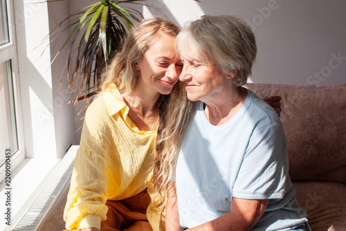 Young woman and elderly woman share joyful embrace, smiling with faces close together. Loving family members sit beside sunlit window, showing warmth and companionship, seated close together on sofa.