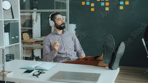 Obraz na plátně Arab guy is having fun in modern office listening to music pretending to play the guitar wearing wireless headphones relaxing at desk alone
