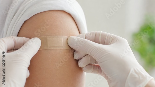 Close up of gloved hands putting adhesive bandage on arm after vaccination in clinic